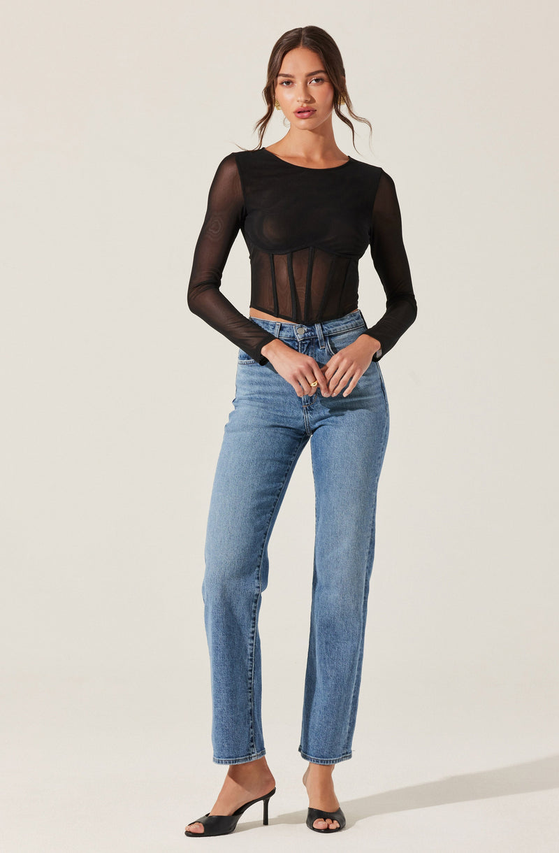 A woman is wearing a sheer black long-sleeve top with a round neckline. The top has a fitted bodice with vertical boning detail and is made of see-through fabric, revealing her skin underneath. She pairs this with high-waisted, straight-leg blue jeans and black open-toe high-heeled shoes.