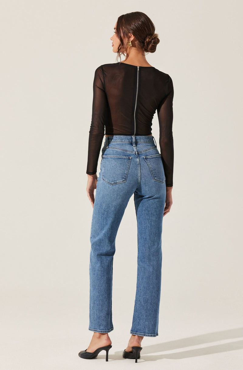 The image shows a woman wearing a fitted, long-sleeved black top made of sheer material, with a visible zipper running down the back. She is also wearing high-waisted, light blue jeans with a relaxed fit and classic five-pocket styling. She has on black high-heeled shoes and gold earrings. Her hair is styled in a low bun.