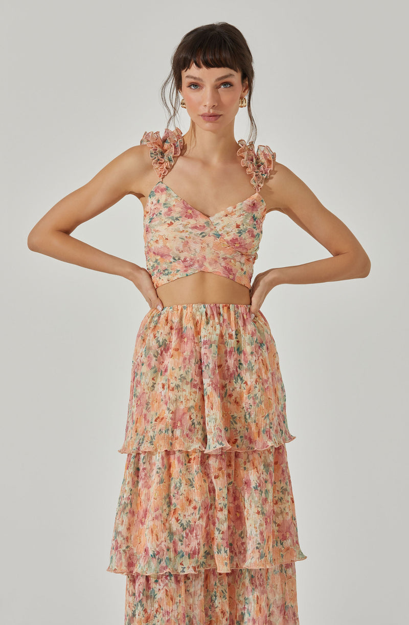 A woman is wearing a sleeveless, floral-patterned crop top with ruffled shoulder straps and a sweetheart neckline. She is also dressed in a high-waisted, tiered, flowy skirt made of lightweight fabric with a matching floral print.