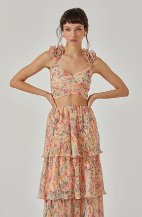 A woman is wearing a sleeveless, floral-patterned crop top with ruffled shoulder straps and a sweetheart neckline. She is also dressed in a high-waisted, tiered, flowy skirt made of lightweight fabric with a matching floral print.