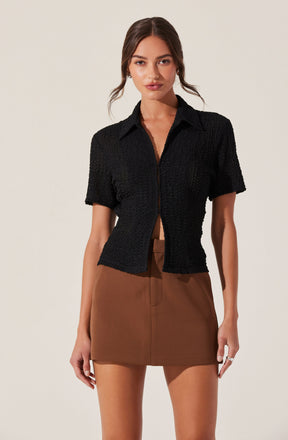 A woman is wearing a short-sleeved, black textured button-up shirt with a collar. The shirt is slightly cropped, ending above the waist. She pairs it with a high-waisted, brown mini skirt with a front zipper detail.