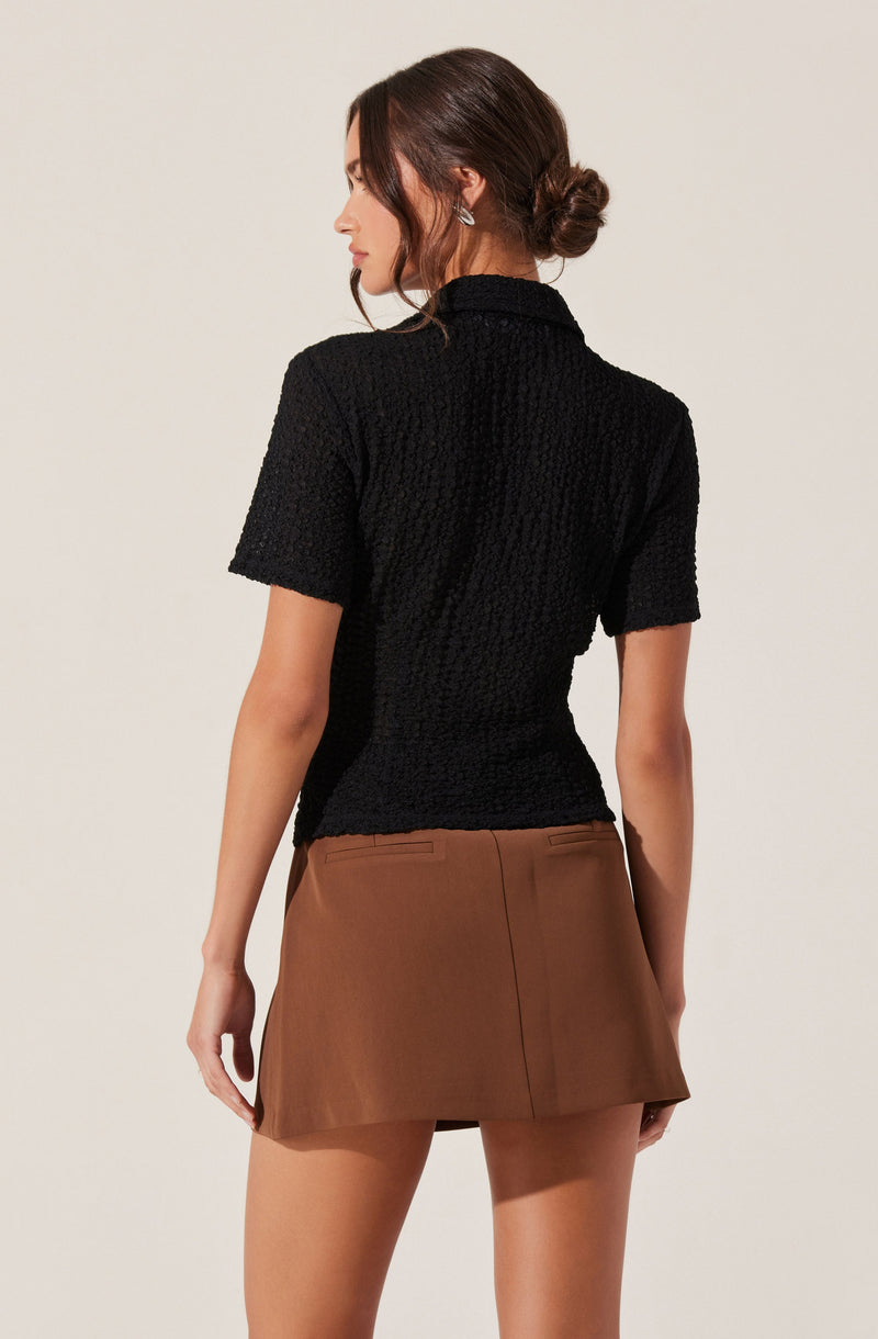 A woman is wearing a black textured short-sleeve top with a high collar, paired with a brown mini skirt.