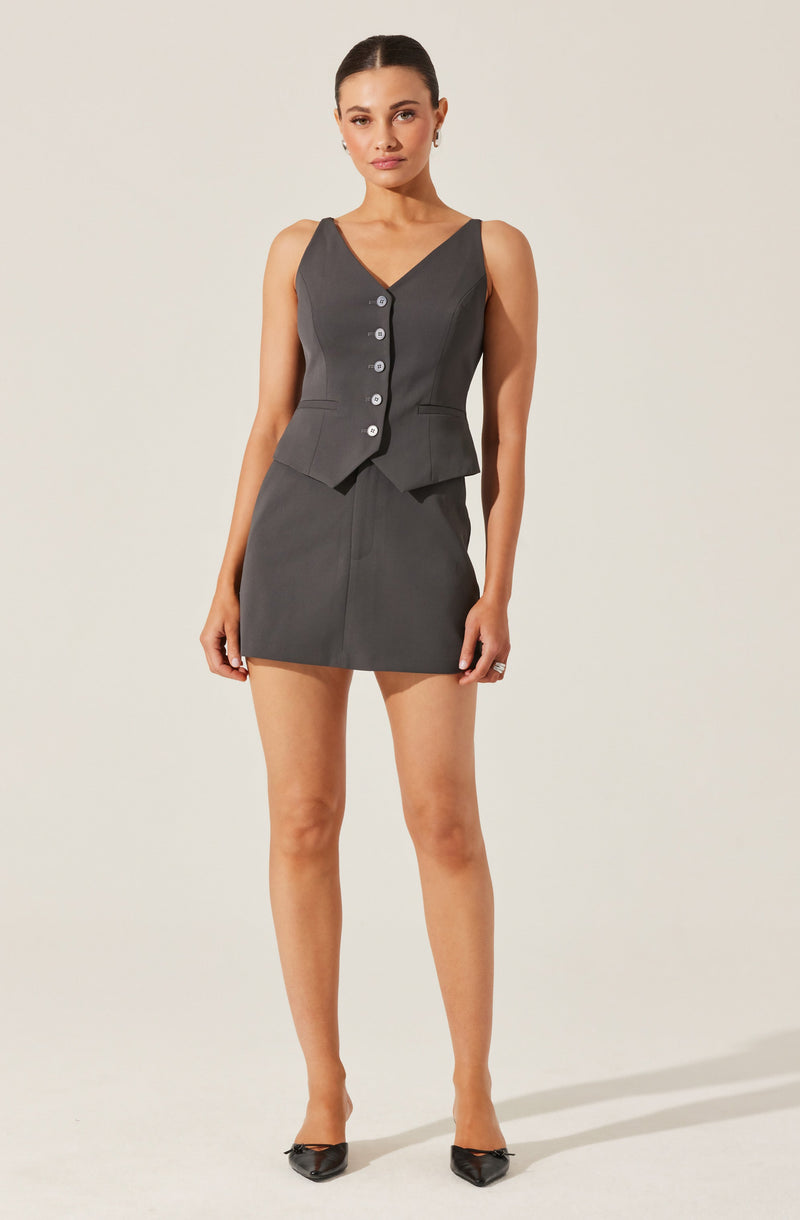 A woman is wearing a sleeveless, dark gray vest with a V-neckline and a button-down front, paired with a matching gray skirt. She is also wearing black pointed-toe heels with embellishments.