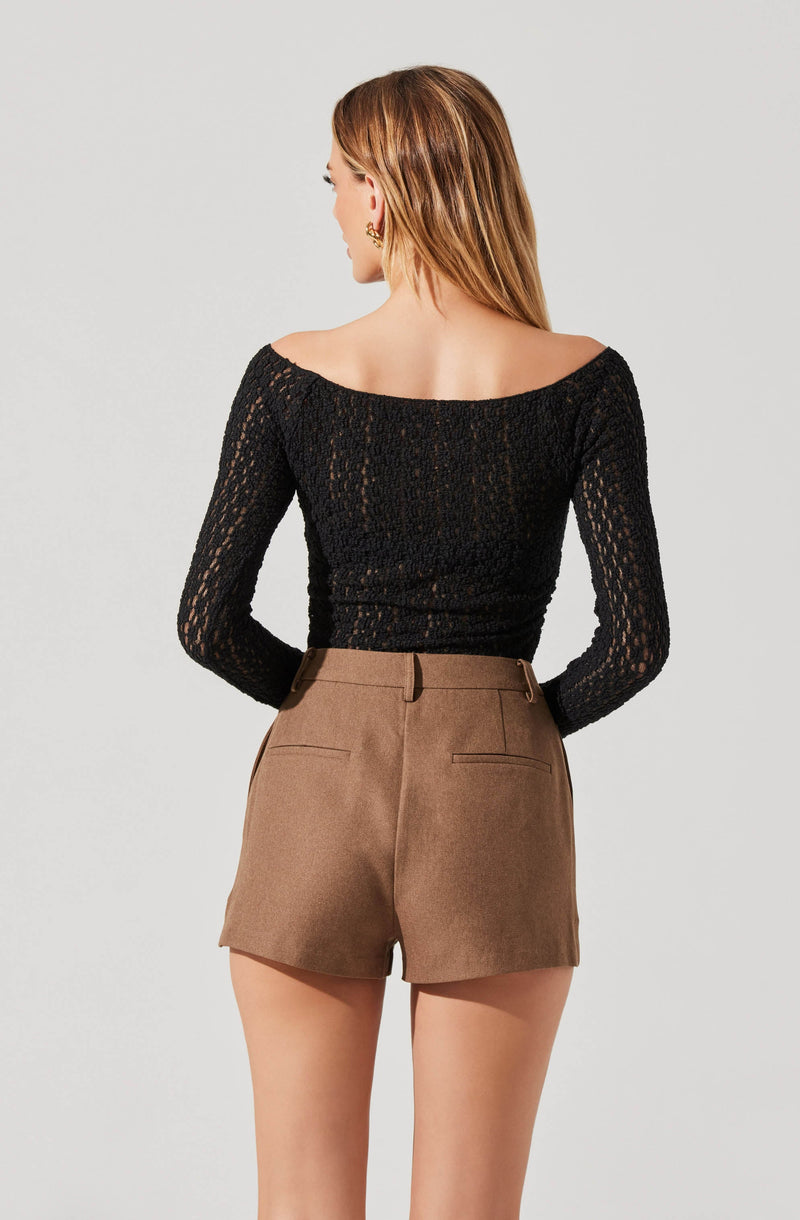 A woman is wearing a black, long-sleeved, off-the-shoulder top made of a lace or crochet material, paired with high-waisted brown shorts that have belt loops and back welt pockets.
