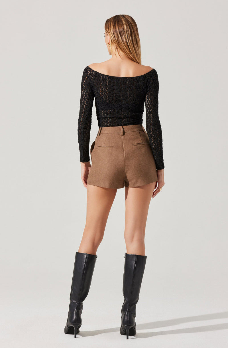 A woman is wearing a black, long-sleeved, off-the-shoulder knitted top paired with high-waisted, khaki-colored shorts. She is also wearing tall black leather boots with high heels.