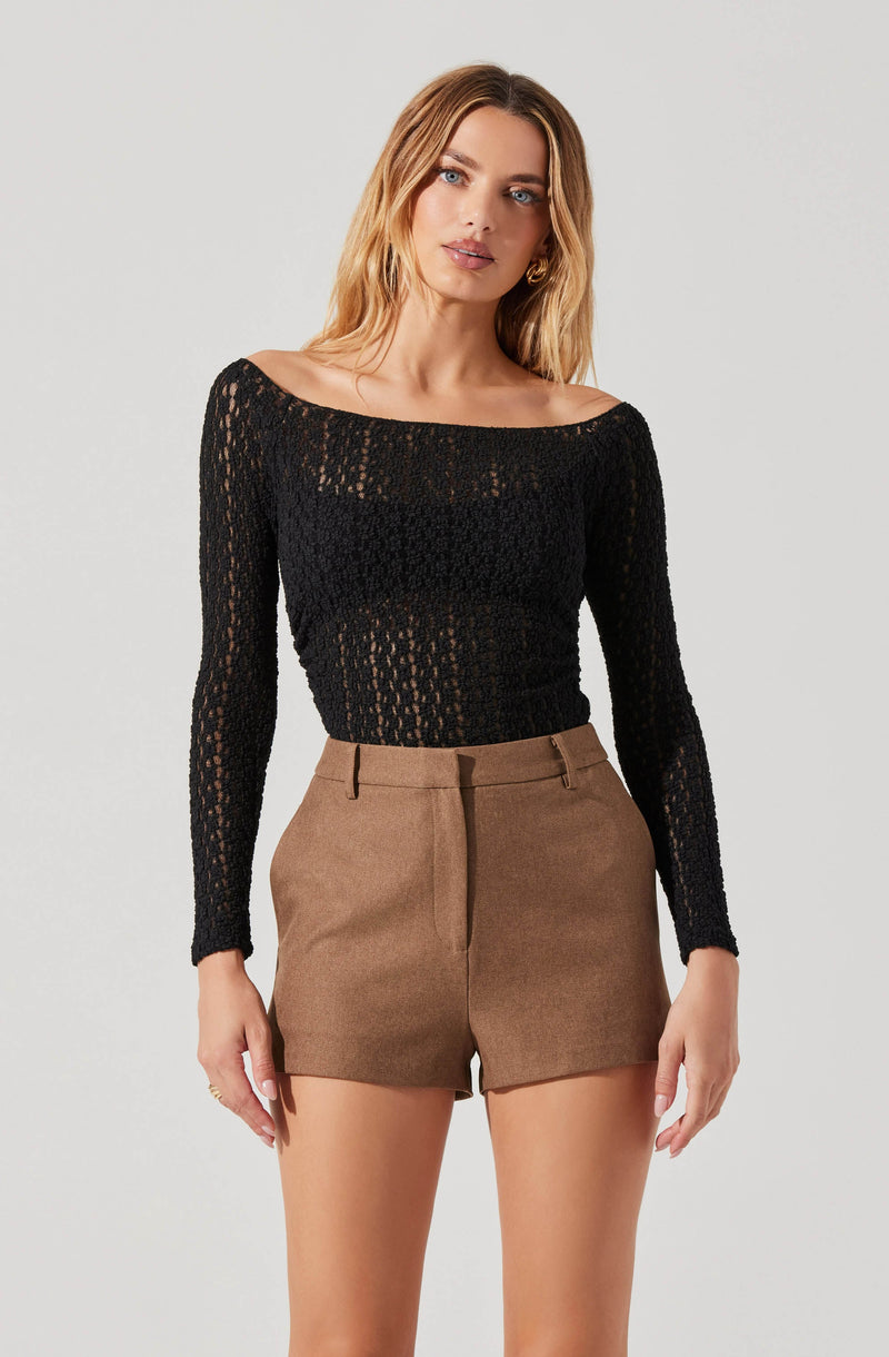 A woman is wearing a black, long-sleeved, open-knit, scoop-neck top paired with high-waisted, tailored brown shorts.