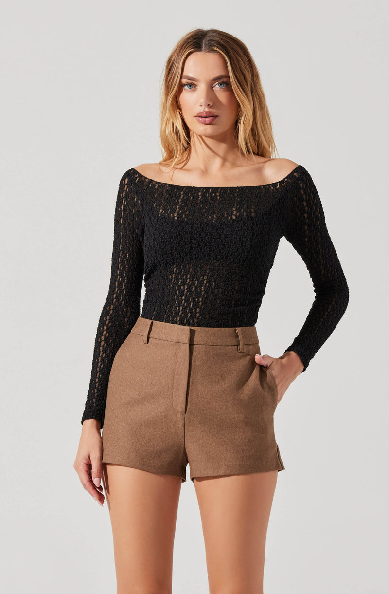 A woman is wearing a black, long-sleeved, off-the-shoulder top made of a open-knit, textured fabric. She pairs it with high-waisted, tailored brown shorts with belt loops.
