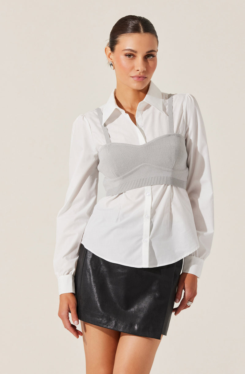 A woman is wearing a white button-up shirt with long sleeves and a collar. Over the shirt, she has a light gray, cropped knit bustier-style top with spaghetti straps. She is also wearing a black, shiny leather mini skirt.