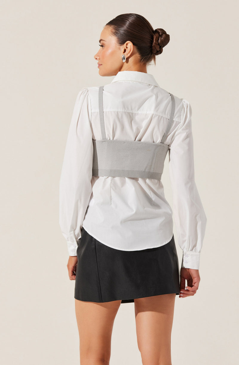 A woman is wearing a white long-sleeved button-up shirt with a collar. Over the shirt, she has a light gray sleeveless knit crop top with thin straps. She is also wearing a black mini skirt. Her hair is styled in a sleek, low bun.
