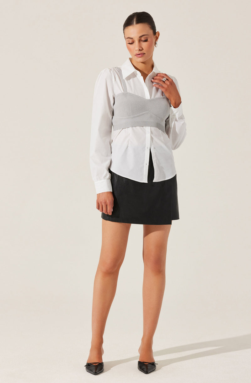 A woman is wearing a white button-up shirt with long sleeves and a collar. Over the shirt, she has layered a gray cropped knit bustier top. She is also wearing a black mini skirt and pointed black shoes.
