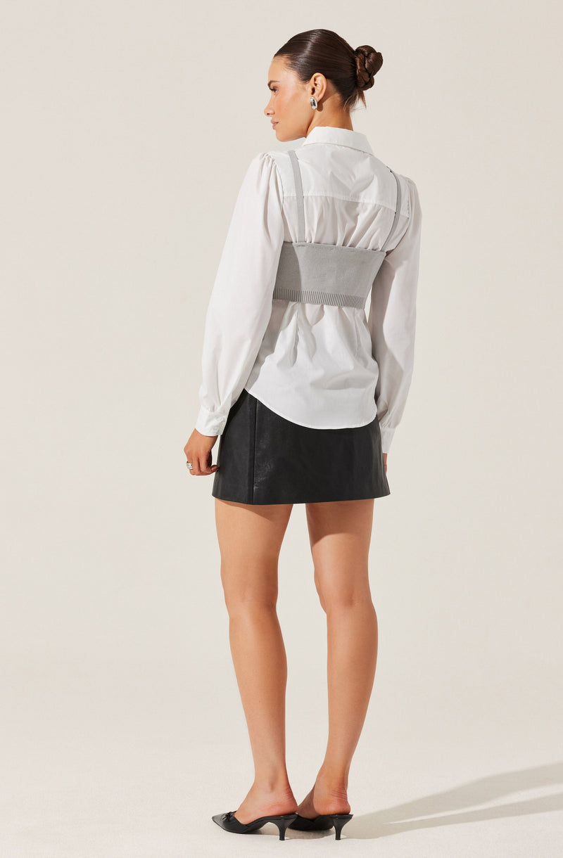 A woman is wearing a white long-sleeve button-up shirt with puffed sleeves and a collar. Over the shirt, she has on a light gray waist cincher or belt. She is also dressed in a black mini skirt made of a leather-like material and is wearing black open-toe high heels.