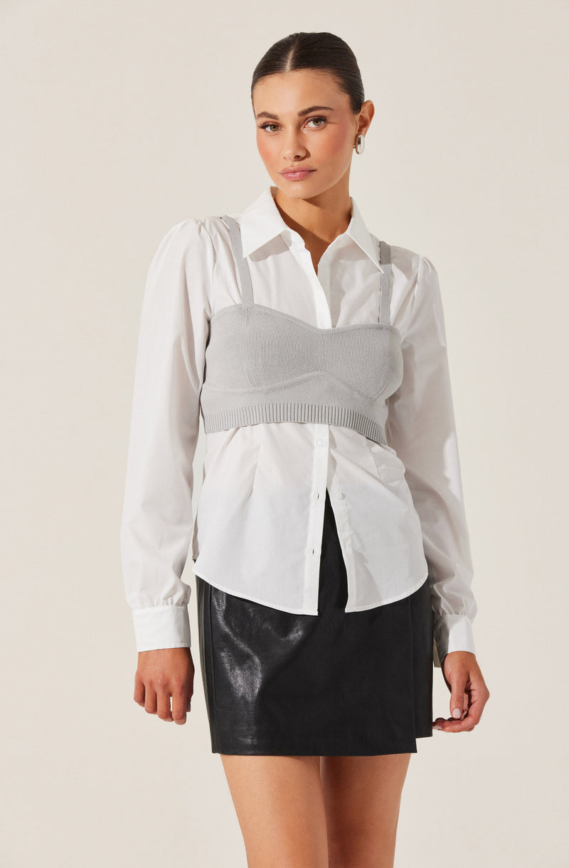 A woman is wearing a white button-up shirt with long sleeves and a classic collar. Over the shirt, she has on a light gray knitted bustier-style crop top with wide shoulder straps. She pairs this with a black, shiny, leather-look mini skirt.