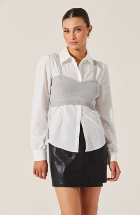 A woman is wearing a white button-up shirt with long sleeves and a classic collar. Over the shirt, she has on a light gray knitted bustier-style crop top with wide shoulder straps. She pairs this with a black, shiny, leather-look mini skirt.