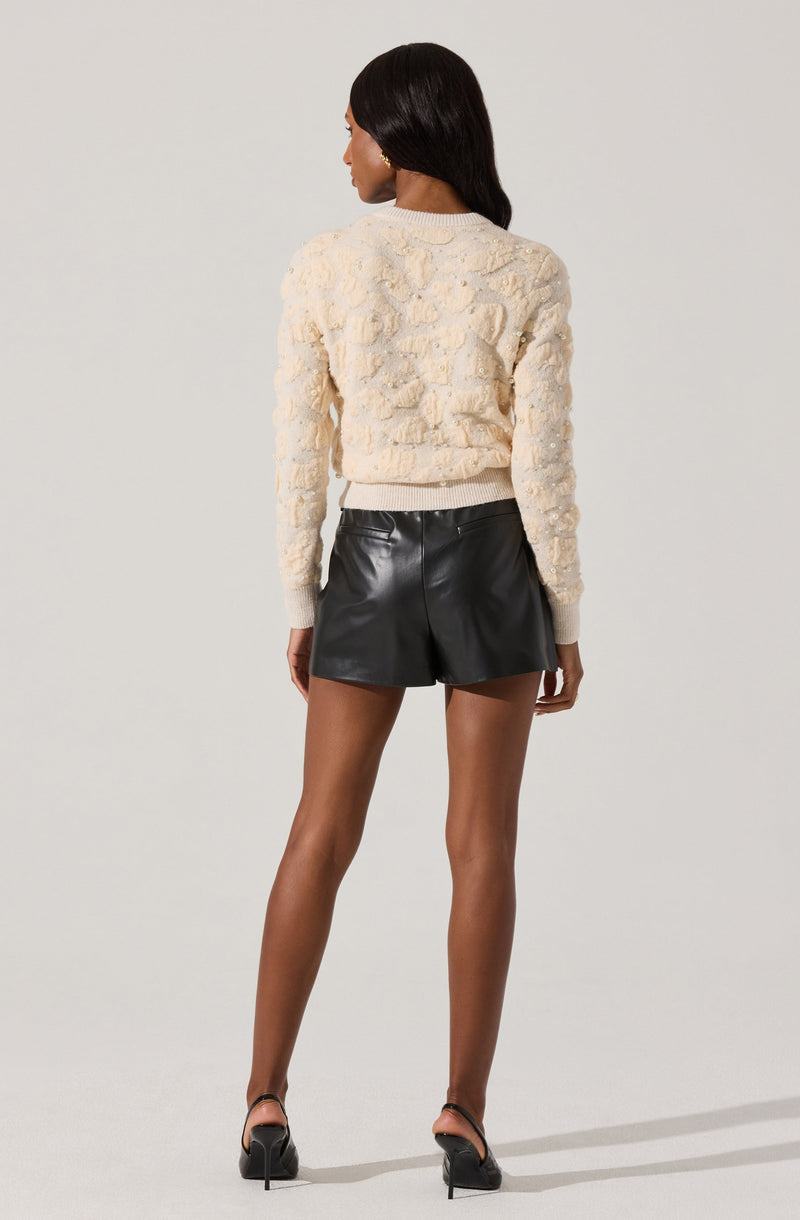 A woman is wearing a cream-colored, textured sweater with a 3D floral pattern, paired with high-waisted black leather shorts and black high-heeled shoes.