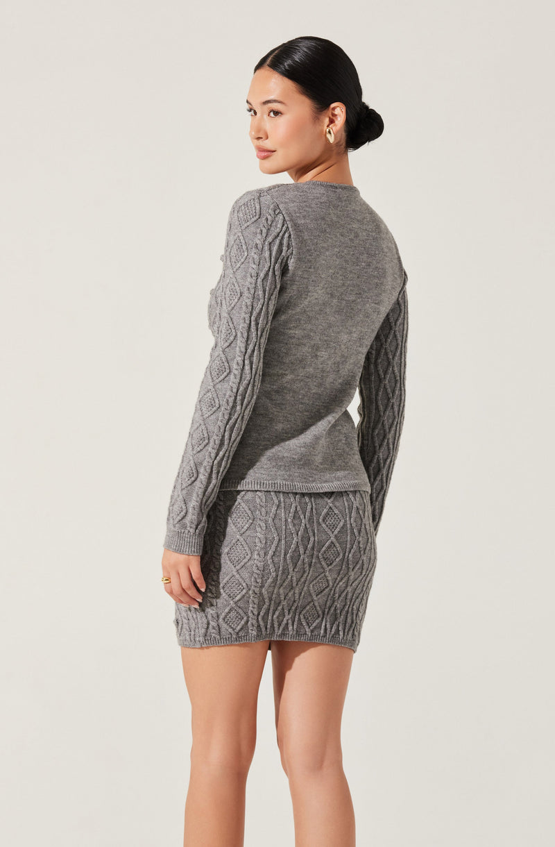 A woman is wearing a matching gray knit sweater and skirt set. The sweater features a solid dark gray front and textured patterned sleeves with a diamond and cable knit design. The skirt is a short, fitted style with the same diamond and cable knit pattern as the sleeves on the sweater. The woman has dark hair styled in a low bun and is accessorizing with small earrings.