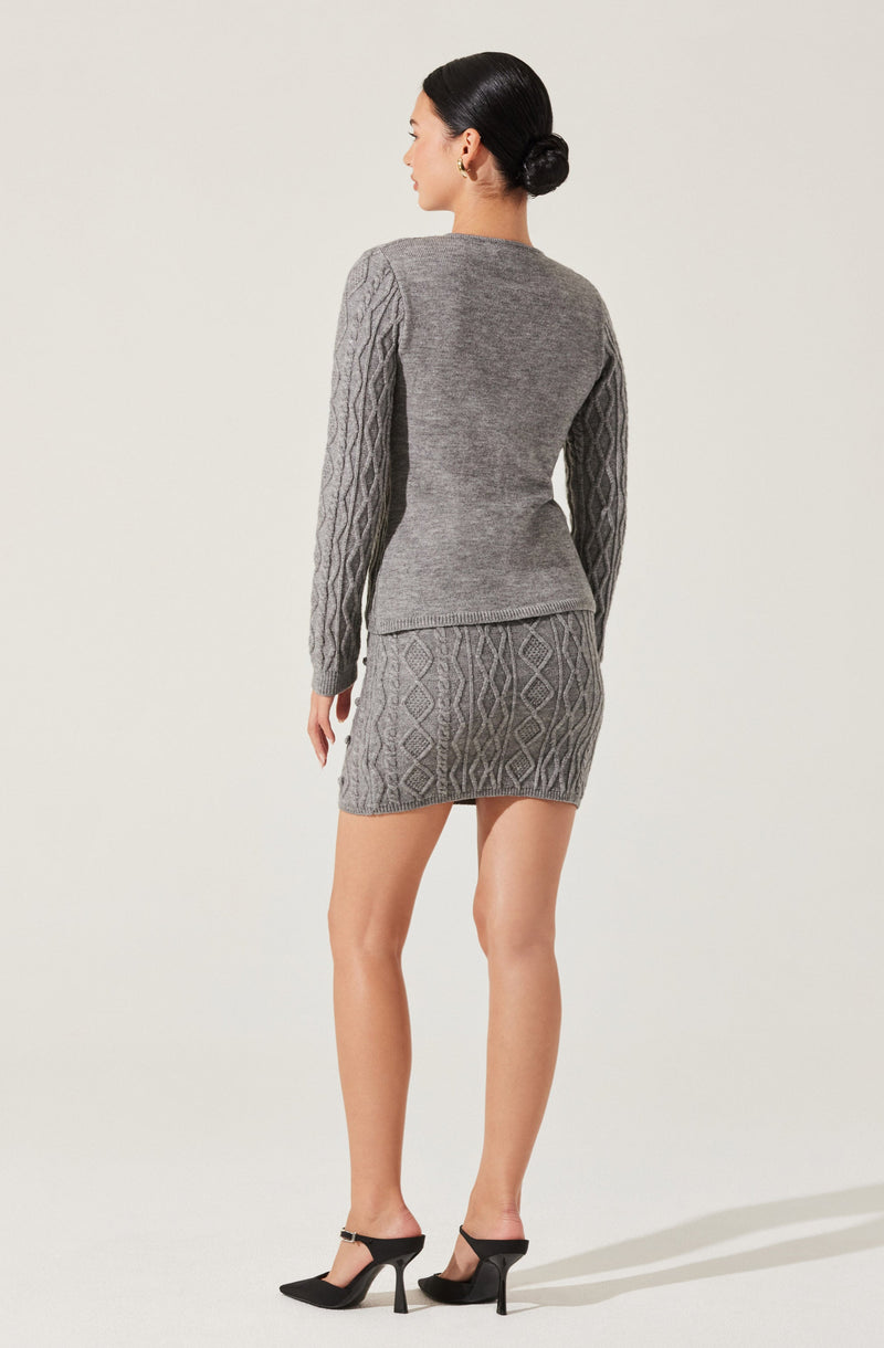 A woman is wearing a matching gray knit sweater and skirt set. The sweater has long sleeves with a diamond cable knit pattern on the sleeves and a plain knit body. The skirt features a diamond cable knit pattern throughout. She is also wearing black high-heeled shoes.
