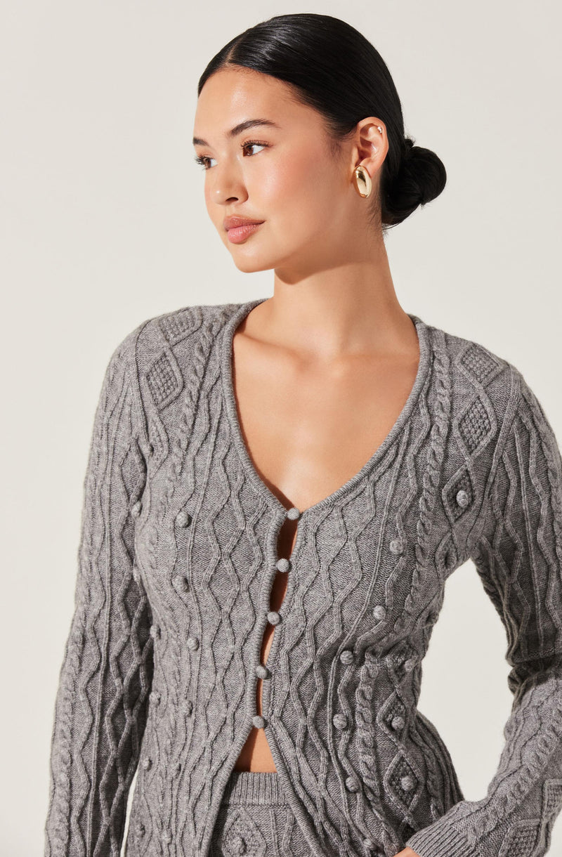 A woman is wearing a gray, long-sleeved, cable-knit cardigan with intricate diamond and braid patterns. The cardigan has a V-neckline with decorative buttons running down the front. She is also wearing gold hoop earrings.