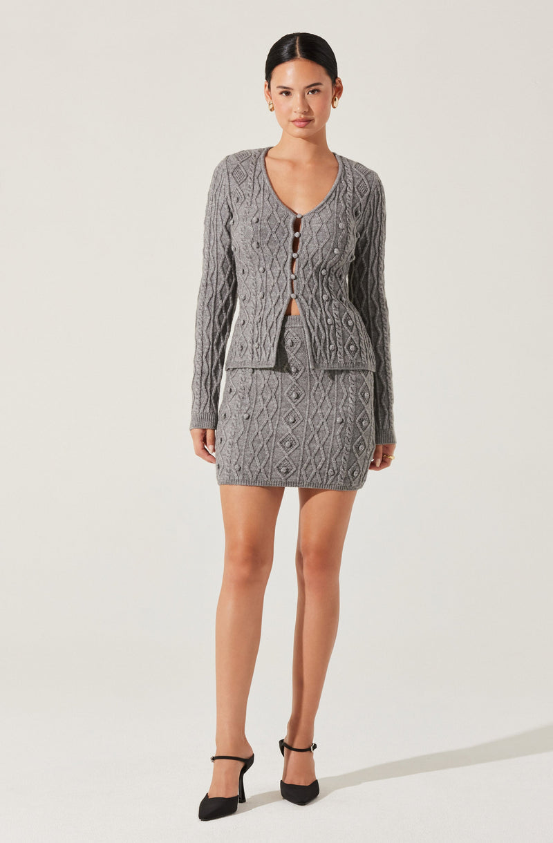 A woman is wearing a matching two-piece outfit consisting of a gray cable-knit cardigan with diamond patterns and a matching gray mini skirt. The cardigan has long sleeves, a round neckline with a button-up front featuring brown buttons, and is slightly cropped. The skirt is fitted and short, ending above the knees.