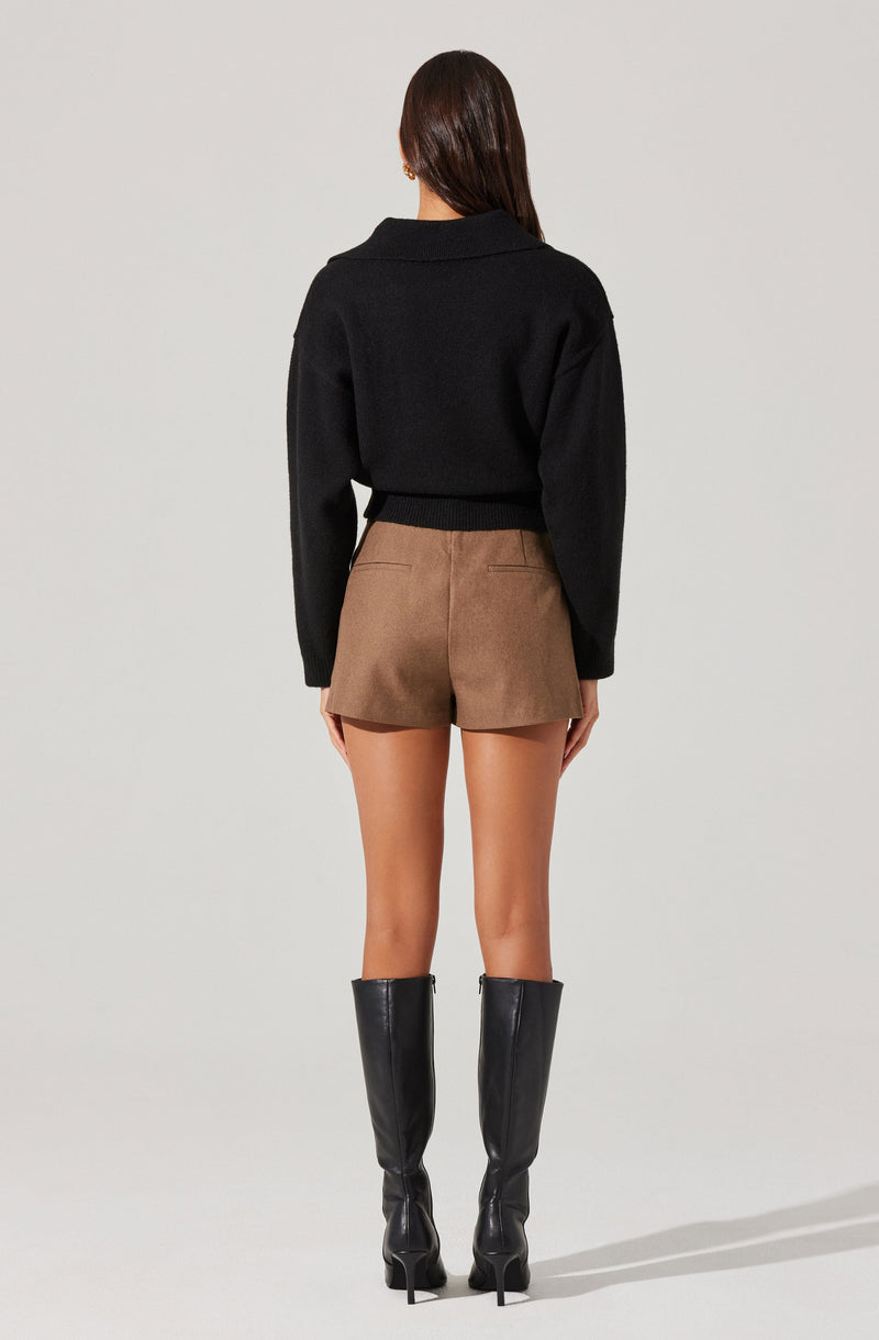 A woman is wearing a black, long-sleeved, cropped sweater with a wide collar, paired with high-waisted, brown shorts. She is also wearing tall, black knee-high boots with high heels.