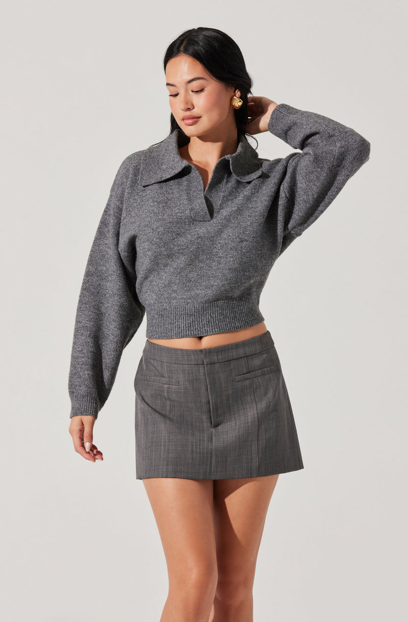 A woman is wearing a cropped, long-sleeved, gray sweater with a collar and partial button placket. She pairs it with a gray, high-waisted, plaid mini skirt that features two front pockets.