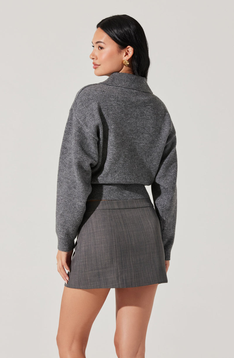 A woman is wearing a gray, cropped, wool-blend cardigan with long sleeves, ribbed cuffs, and a ribbed waistband. She pairs it with a high-waisted, gray plaid mini skirt.