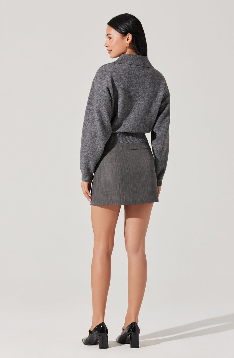 A woman is wearing a dark gray, long-sleeved, collared sweater with ribbed cuffs. She pairs it with a gray plaid mini skirt that has a subtle checkered pattern, and black high-heeled shoes with block heels.