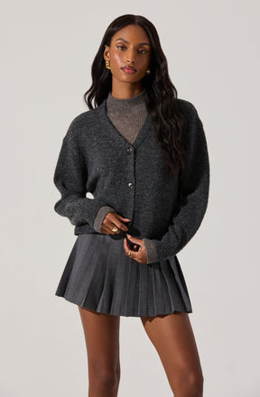 A woman is wearing a dark gray, oversized, button-up cardigan with a textured knit pattern. Underneath, she has a high-neck, taupe-colored sweater. She is also wearing a gray pleated mini skirt with vertical stripes.