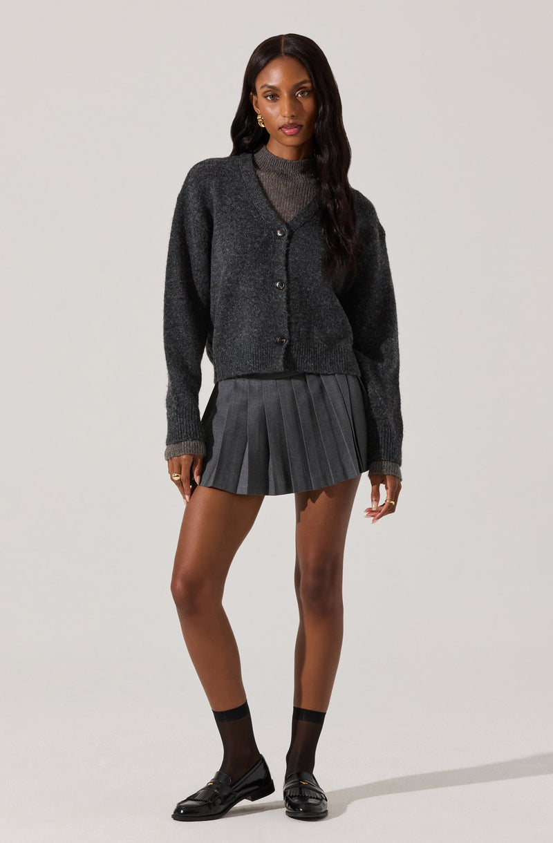 A young woman is wearing a dark gray, oversized button-up cardigan over a high-neck, long-sleeve sweater in a similar gray tone. She has on a pleated, dark gray skirt that reaches mid-thigh and black ankle socks paired with shiny black loafers. She has long, wavy dark hair and is wearing large gold earrings.