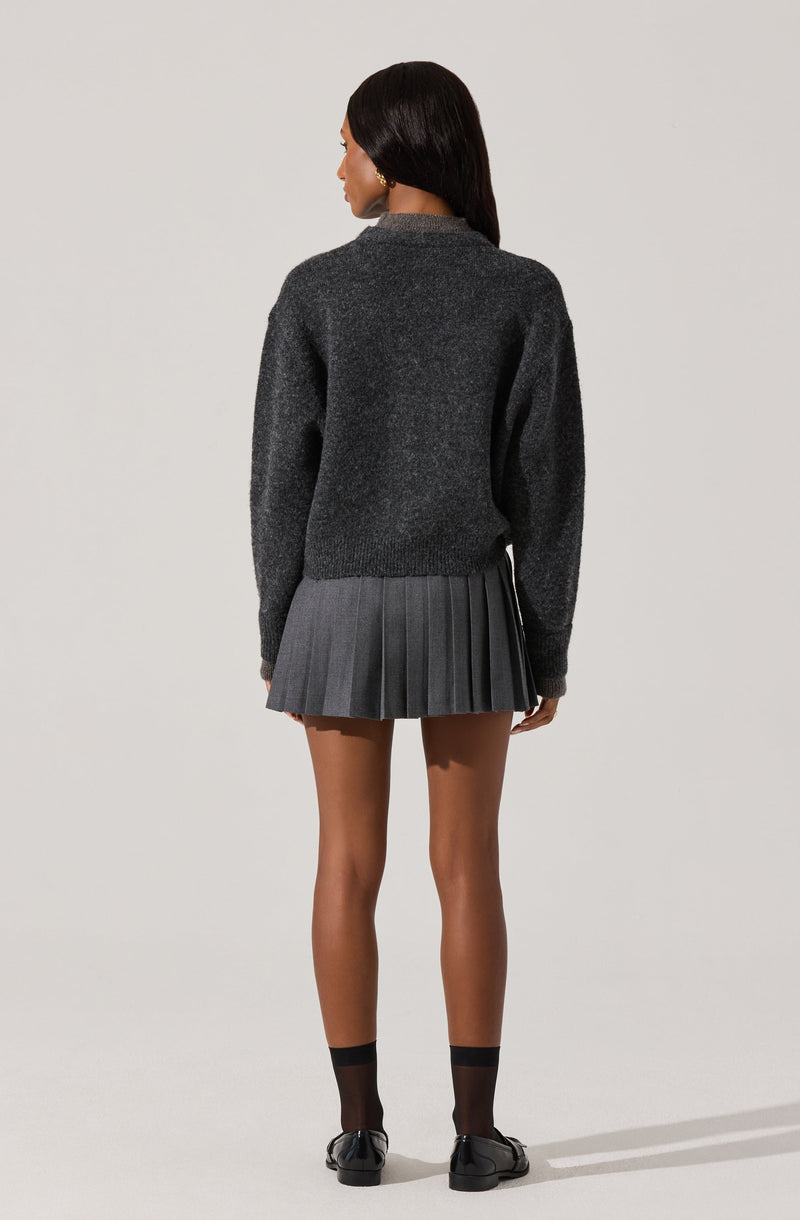 A woman is wearing a dark gray, long-sleeved, crewneck sweater that appears soft and cozy. She is also wearing a dark gray pleated skirt that falls above her knees, with evenly spaced pleats. Her outfit is completed with black ankle socks and black shoes.