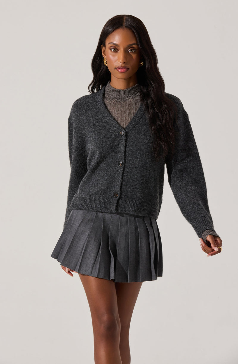 A woman is wearing a dark gray, button-up cardigan sweater over a matching gray knit top. She has on a pleated gray skirt with vertical stripes, and she is accessorized with gold hoop earrings.