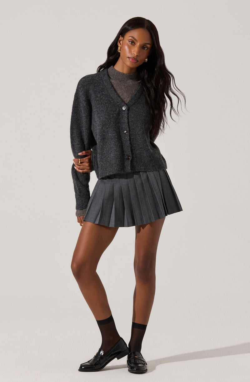 A woman is wearing a dark gray, long-sleeved, button-up cardigan over a matching gray sweater. She has on a pleated, dark gray mini skirt with vertical stripes. She is also wearing sheer black ankle socks and shiny black loafers. She has long, wavy dark hair and is accessorized with gold earrings.