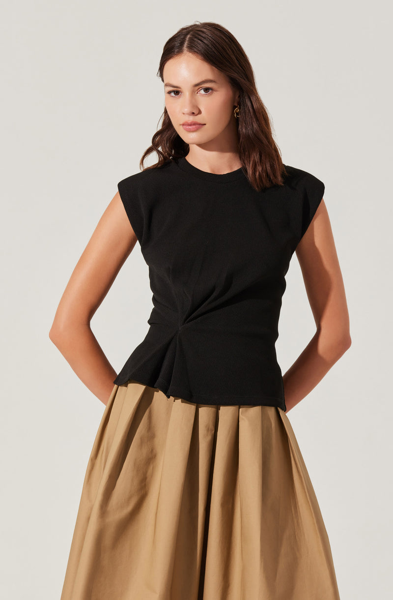 A woman is wearing a sleeveless black top with slightly padded shoulders and a twisted detail at the waist. She is also wearing wide-legged, high-waisted beige pants with pleats.