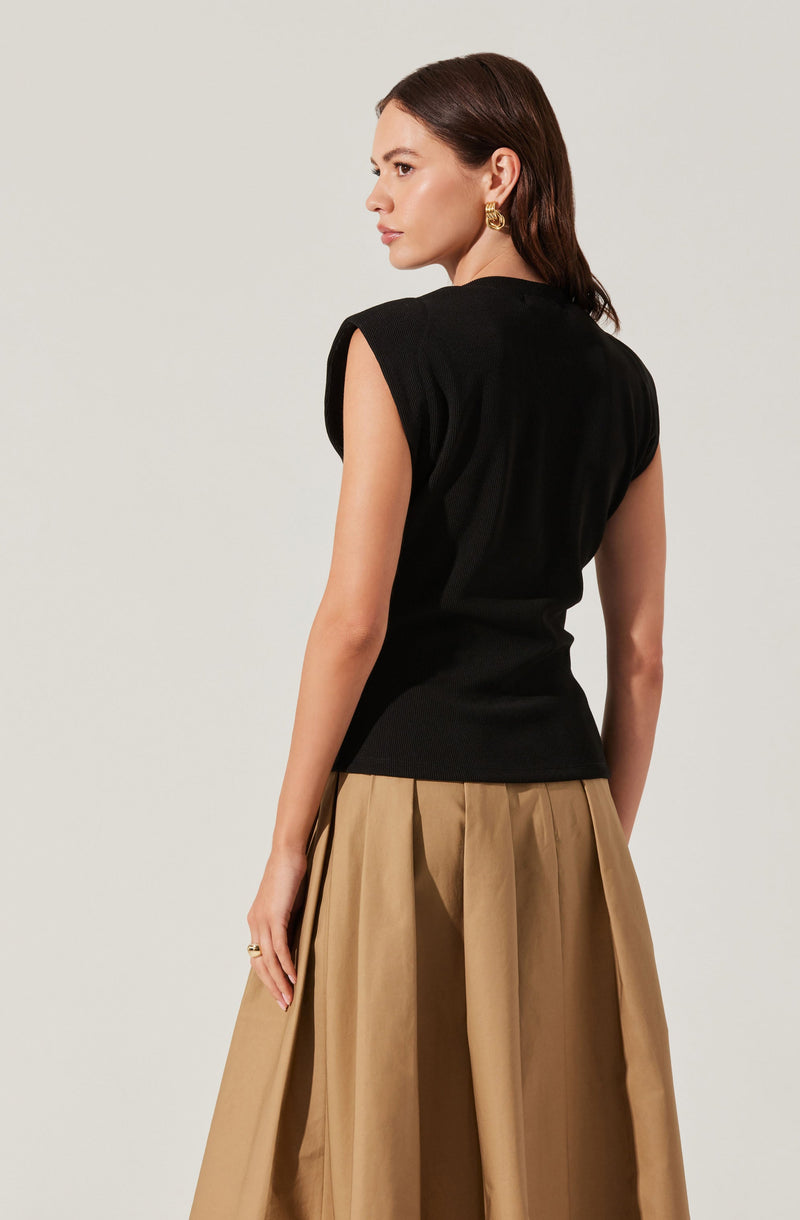 A woman is wearing a sleeveless, black top with padded shoulders and a fitted waist. She pairs it with a high-waisted, full-length beige skirt that has pleats. She also has gold hoop earrings and rings on her fingers.