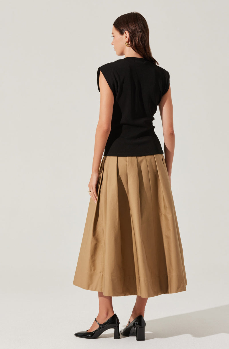 A woman is wearing a black sleeveless top with structured shoulders and a high neckline, paired with a high-waisted, full-length beige pleated skirt that reaches her ankles. She completes the outfit with black ankle-strap high-heeled shoes.