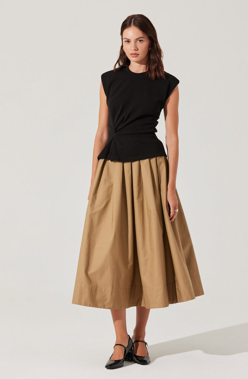 A woman is wearing a sleeveless black top with padded shoulders and a high neckline. She pairs it with a full, beige, pleated midi skirt. She is also wearing black pointed-toe heels with ankle straps.