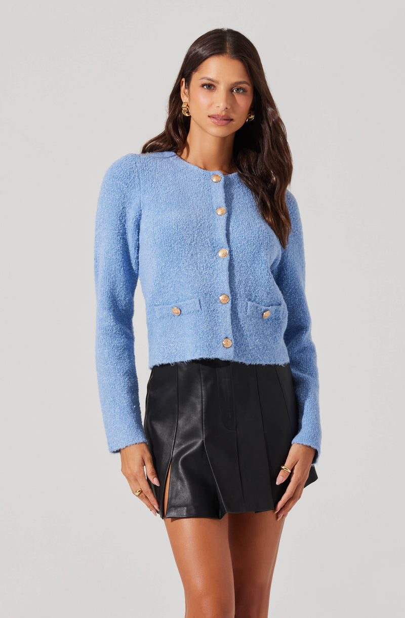 A woman is wearing a light blue textured cardigan with long sleeves and gold buttons down the front. The cardigan has a round neckline and two small pockets at the front. She pairs it with a high-waisted black leather mini skirt with front pleats and side slits.