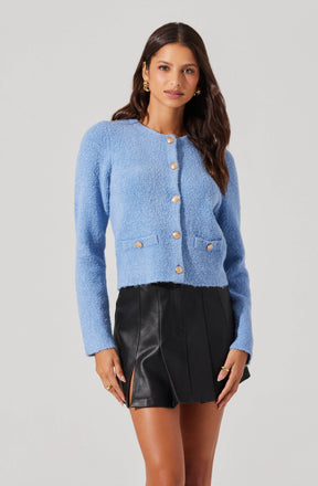 A woman is wearing a light blue textured cardigan with long sleeves and gold buttons down the front. The cardigan has a round neckline and two small pockets at the front. She pairs it with a high-waisted black leather mini skirt with front pleats and side slits.