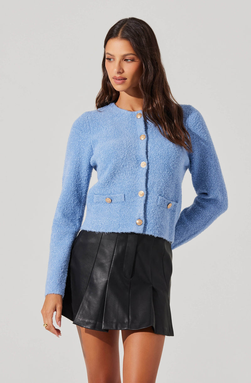 A woman is wearing a bright blue, textured, long-sleeved cardigan with a round neckline and gold buttons down the front. The cardigan has two small, gold-buttoned pockets on the front. She pairs it with a black, high-waisted, pleated leather mini skirt.
