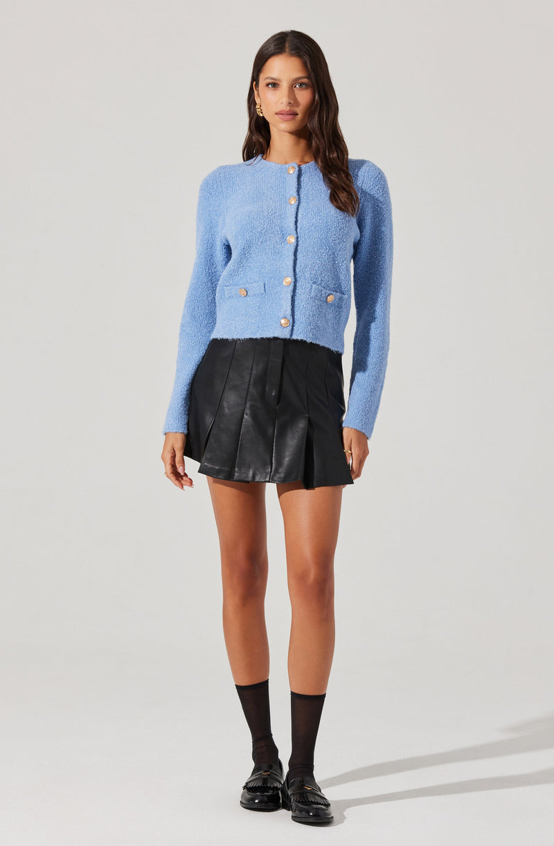 A woman is wearing a light blue, textured, long-sleeve cardigan with pearl buttons down the front and two front pockets with matching pearl buttons. She pairs it with a high-waisted black leather skirt that has pleats. She completes the outfit with black ankle socks and black loafers.