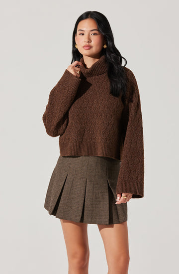 A woman is wearing a cozy, oversized brown turtleneck sweater with long sleeves, paired with a high-waisted, above-the-knee plaid skirt featuring pleats. The outfit has a classic, sophisticated style with earthy tones.