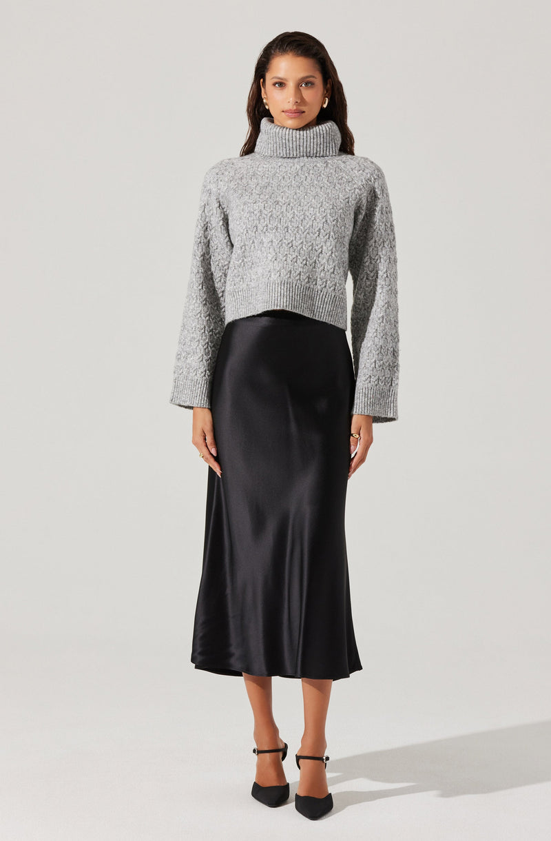 A woman is wearing a light gray, chunky knit turtleneck sweater with long sleeves. She pairs it with a high-waisted, black satin midi skirt that reaches mid-calf. She completes the outfit with black pointed-toe heels with ankle straps.