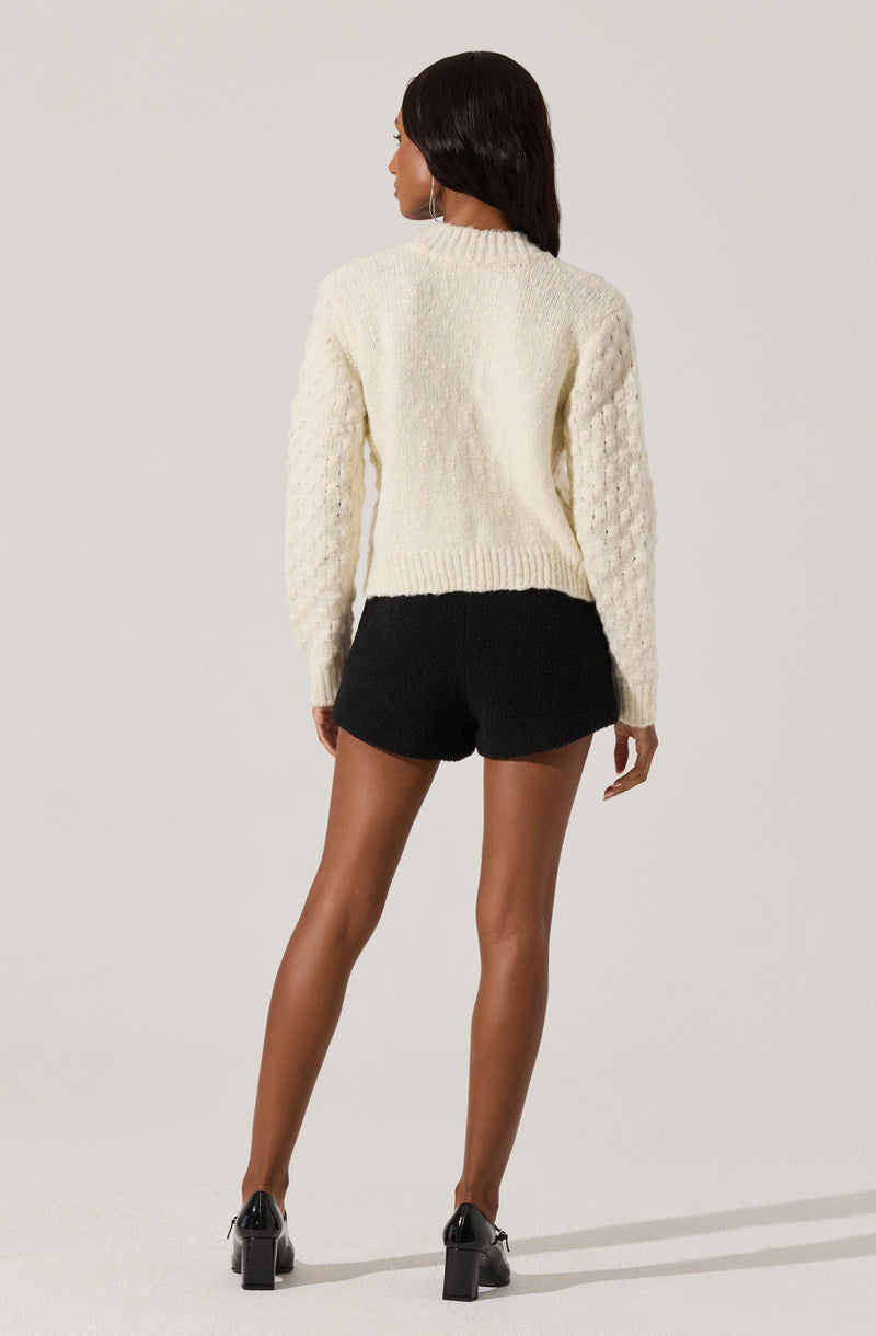 A woman is wearing a chunky, cream-colored knit sweater with textured patterns on the sleeves and a plain front. She pairs it with black shorts and black high-heeled shoes. Her dark hair is styled straight, and she is standing against a plain, light background.