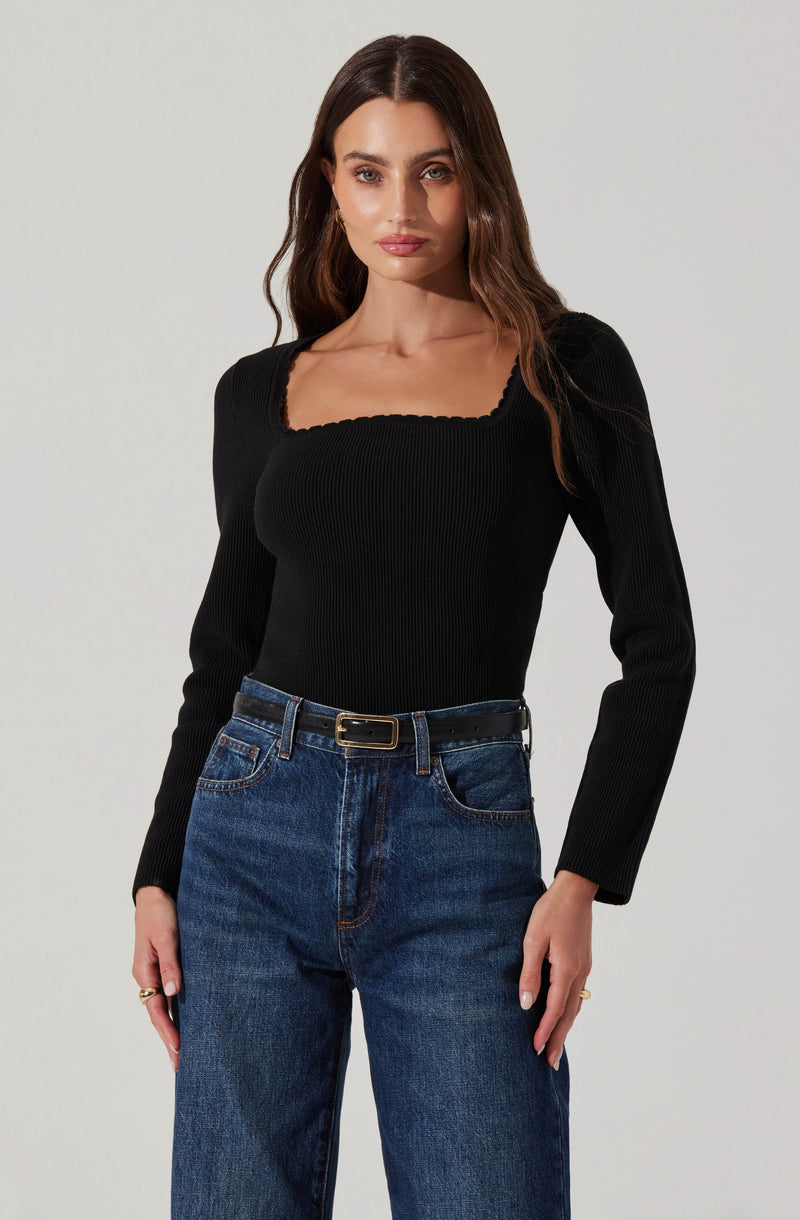 A woman is wearing a fitted black long-sleeve top with a scalloped square neckline and ribbed texture, paired with high-waisted blue jeans with a relaxed fit, secured with a black belt featuring a gold buckle.