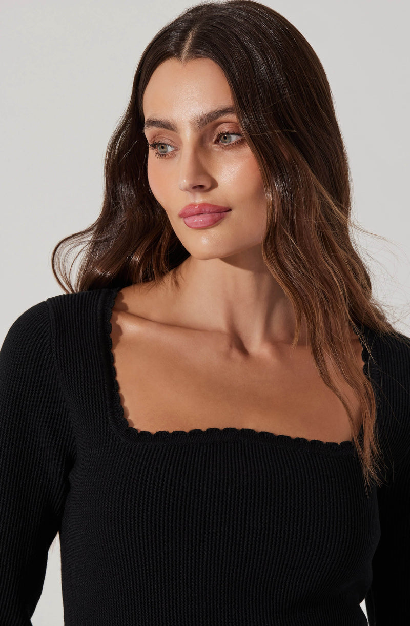 A woman with wavy brown hair is wearing a black, long-sleeved, ribbed top with a square neckline that has a scalloped edge.