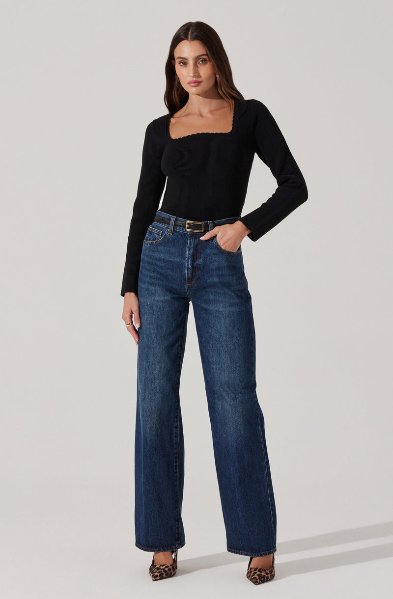 A woman is wearing a fitted black long-sleeve top with a square neckline and scalloped edges, paired with high-waisted, straight-leg blue jeans. She completes the look with a black belt that has a gold buckle and leopard-print high heels.