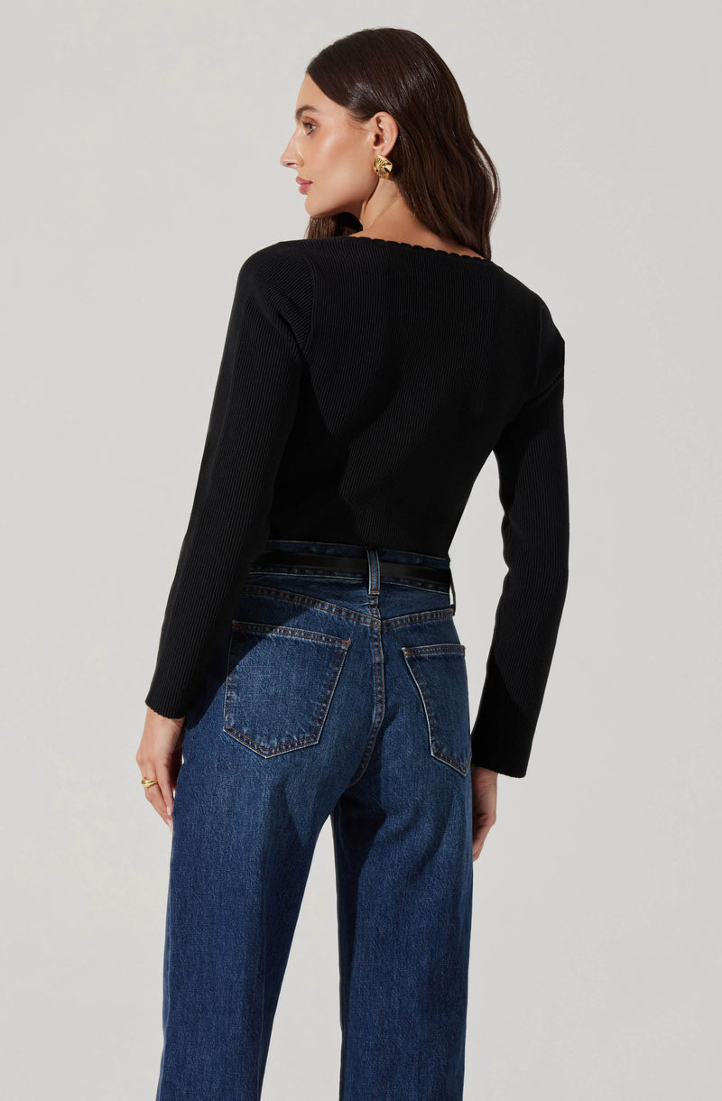 A woman is wearing a fitted black long-sleeve sweater with a subtle ribbed texture and a round neckline. She pairs it with high-waisted blue jeans that have a classic five-pocket design and a slightly relaxed fit. The woman also has on large, gold statement earrings.