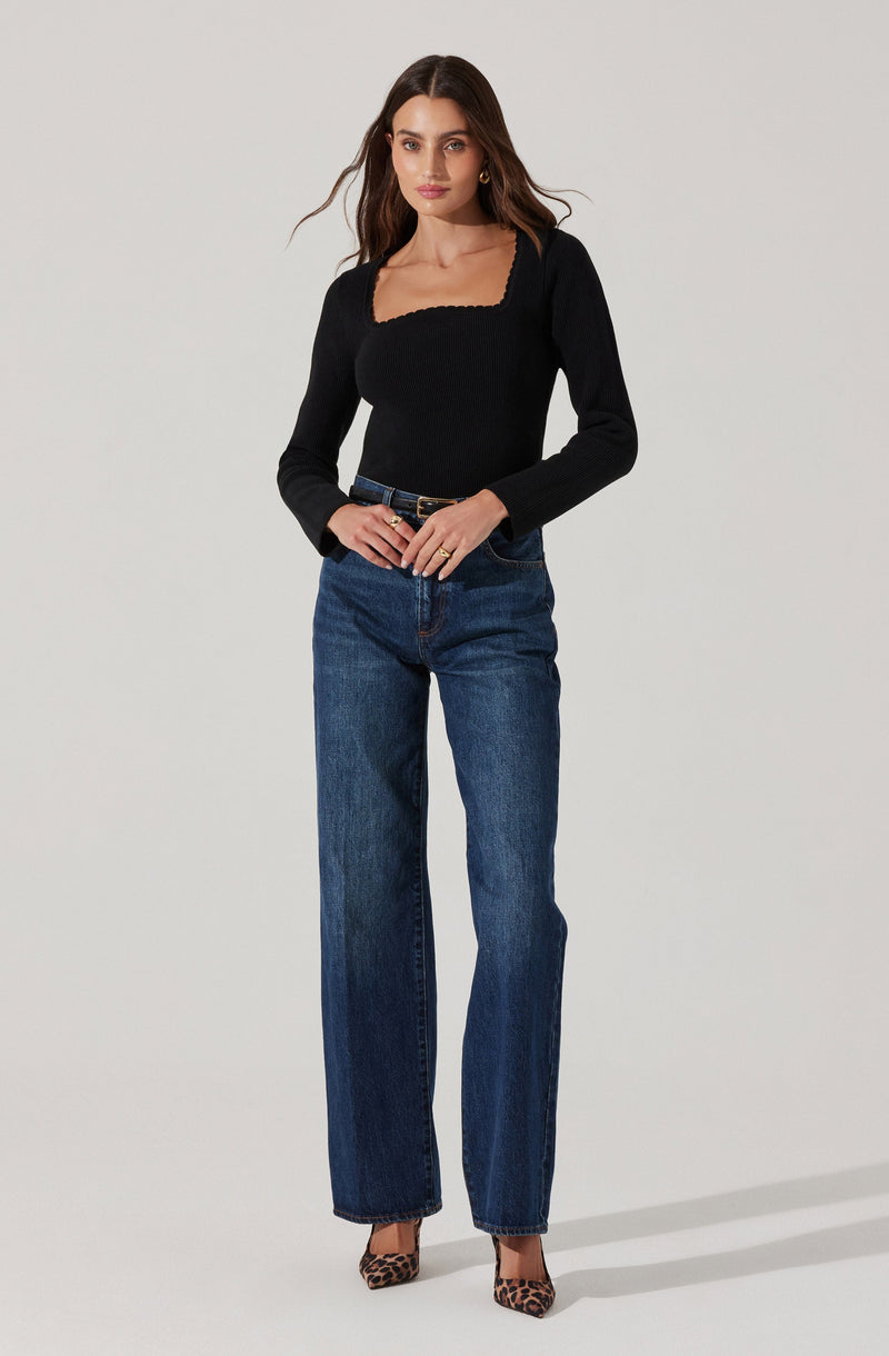 A woman is wearing a fitted black long-sleeve top with a square neckline and a textured hem detail. She pairs it with high-waisted, straight-leg blue jeans and leopard-print pointed-toe heels.