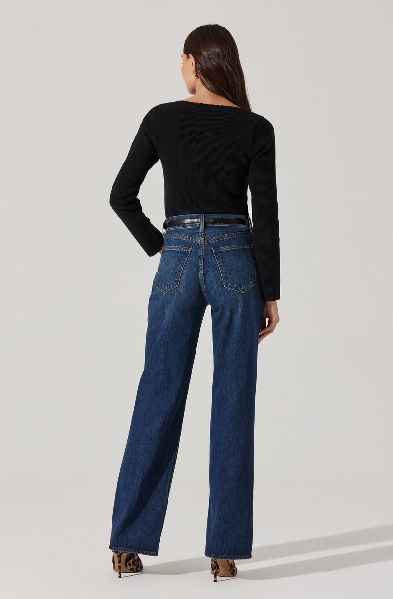 A person is wearing a black long-sleeve top with a slightly frayed neckline, tucked into high-waisted, straight-leg blue jeans. The jeans have prominent back pockets with orange stitching and are paired with a black belt. The individual is wearing animal print high-heeled shoes.