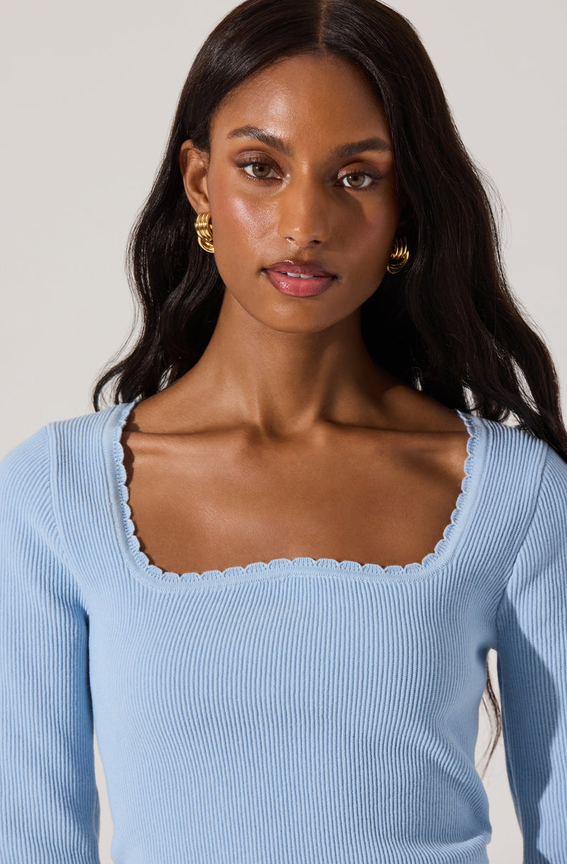 A woman is wearing a light blue, ribbed, long-sleeved top with a square neckline that has scalloped edging. She is accessorized with large, gold hoop earrings.