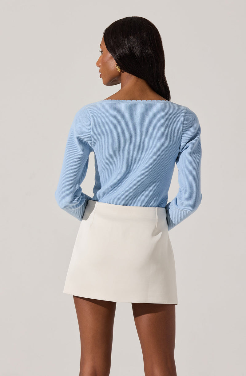 A woman is wearing a light blue, long-sleeved knit sweater with a subtle ribbed texture and a scalloped neckline, paired with a high-waisted white skirt that is short and A-line.